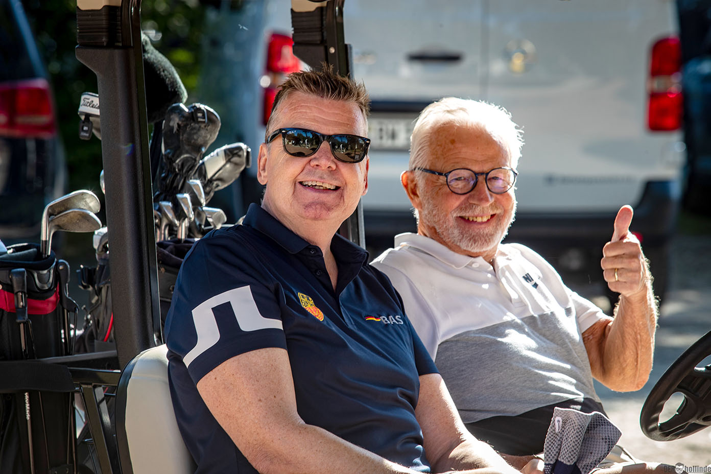 Zwei Erwachsene sitzen in einem Golfwagen, einer trägt eine Sonnenbrille und der andere zeigt einen Daumen nach oben, im Hintergrund sind Golfschläger und Fahrzeuge zu sehen.