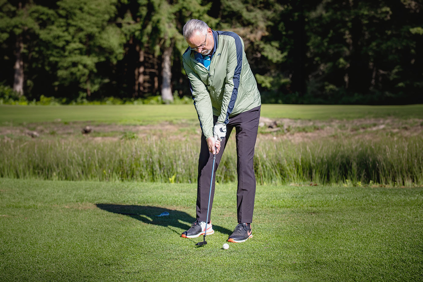 Eine Person mit grauen Haaren trägt eine grüne Jacke und schwarze Hosen und bereitet sich auf einem Golfplatz darauf vor, einen Golfball mit einem Schläger zu schlagen.