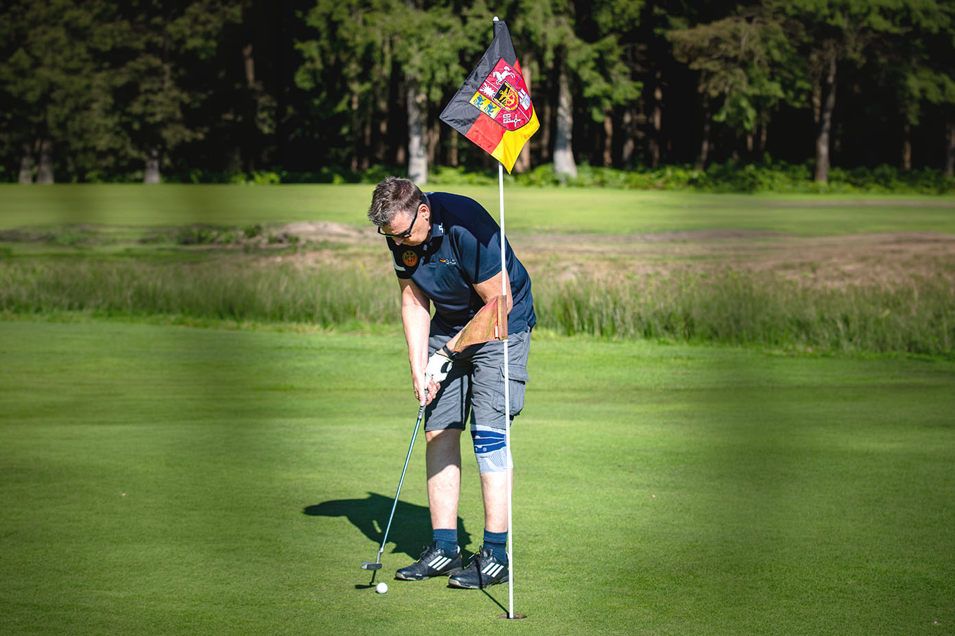 Eine Person mit sportlicher Kleidung und Kniebandage spielt Golf und bereitet sich darauf vor, den Ball in das Loch mit einer Fahne zu schlagen.