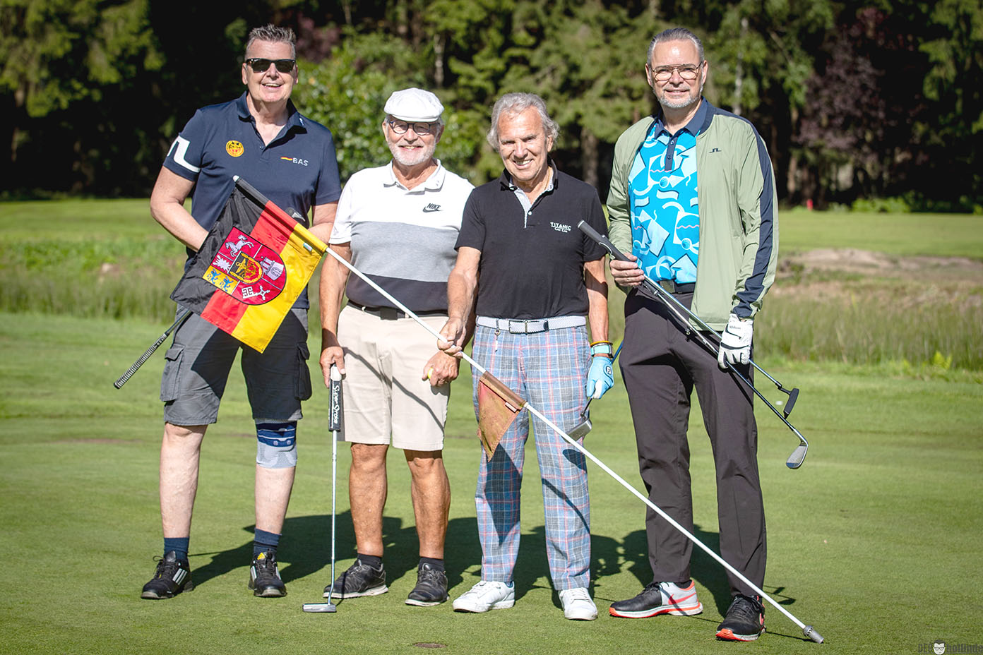 Vier Erwachsene stehen auf einem Golfplatz, zwei halten Golfschläger, einer hält eine Fahne mit den Farben Schwarz, Rot und Gelb, und alle tragen sportliche Kleidung.
