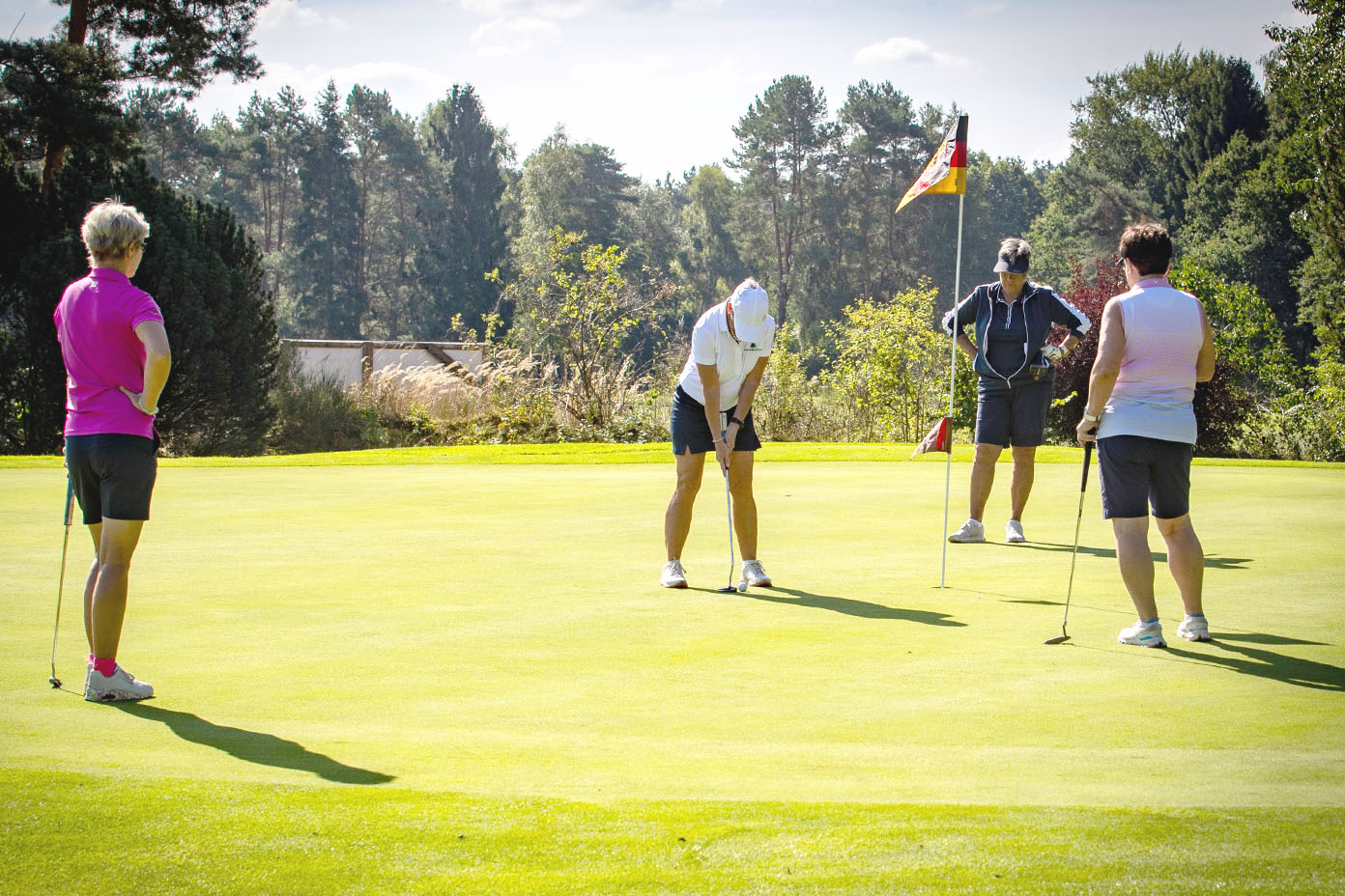 Vier Personen in sportlicher Kleidung spielen bei sonnigem Wetter Golf auf einem grünen Golfplatz mit Bäumen im Hintergrund.