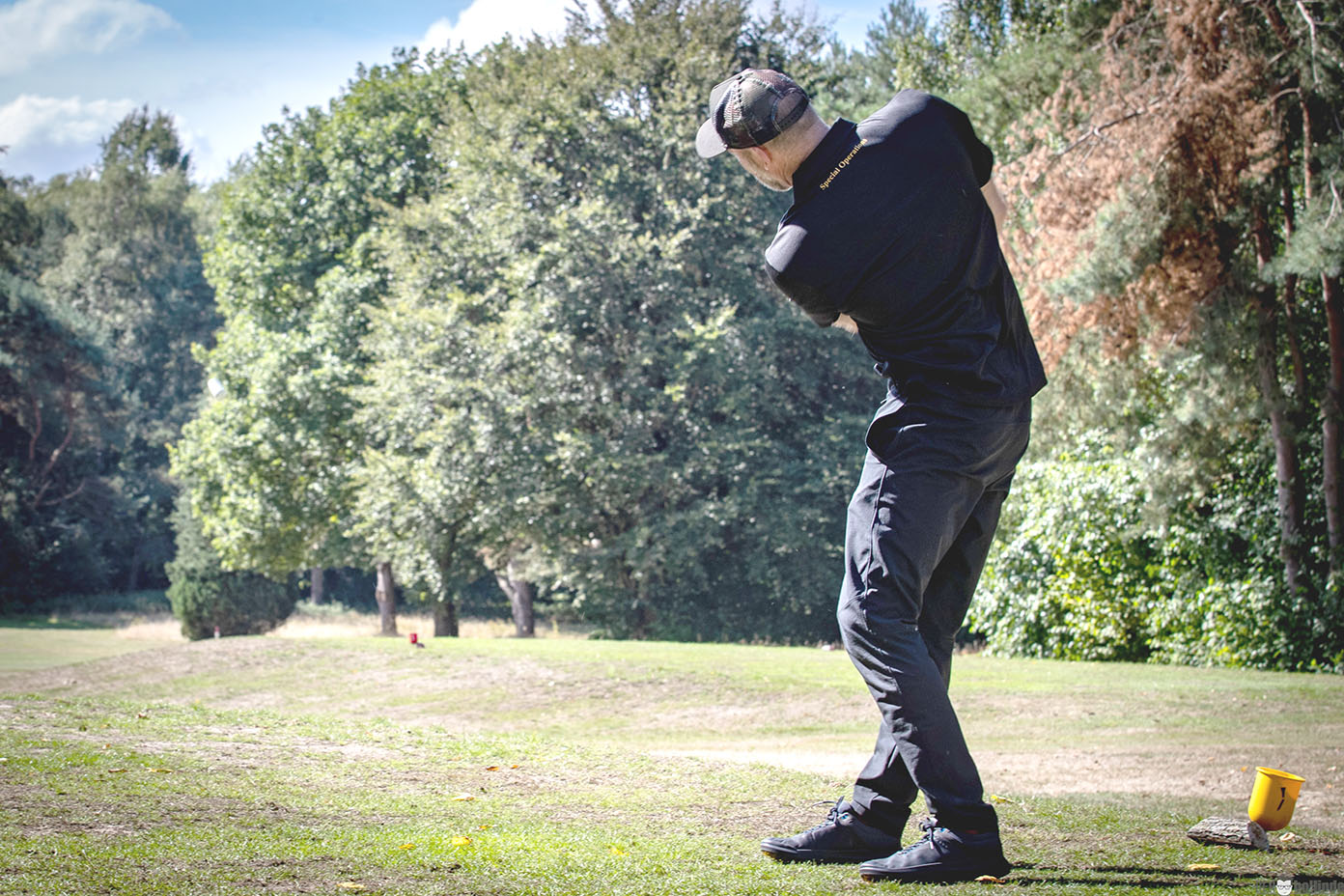 Eine Person in schwarzer Sportkleidung und Baseballkappe schlägt auf einem Golfplatz mit grüner Wiese und Bäumen im Hintergrund einen Golfschläger.