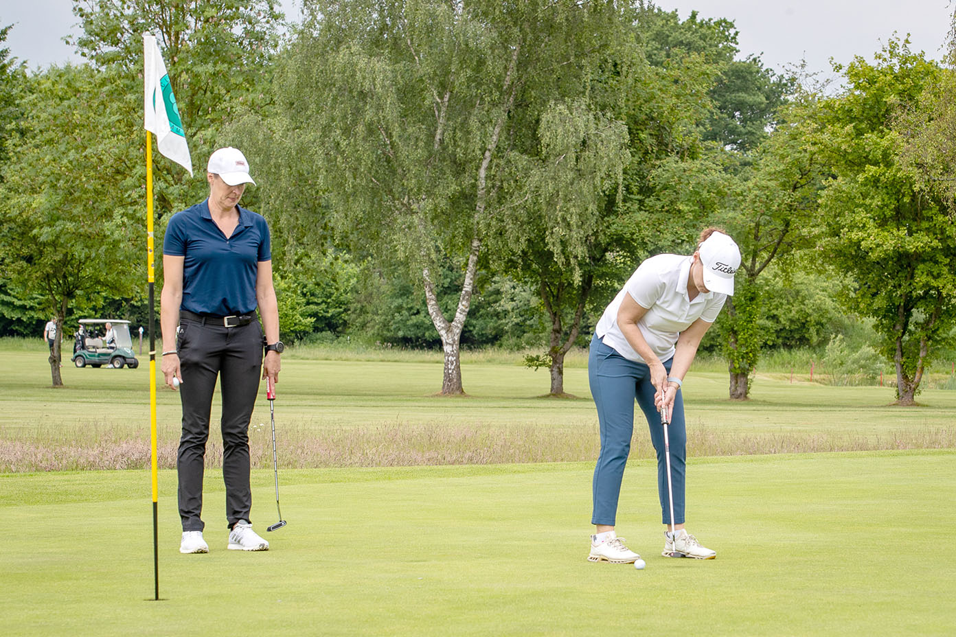 Zwei Personen in sportlicher Kleidung spielen Golf auf einem grünen Golfplatz, wobei eine Person gerade puttet und die andere zuschaut.