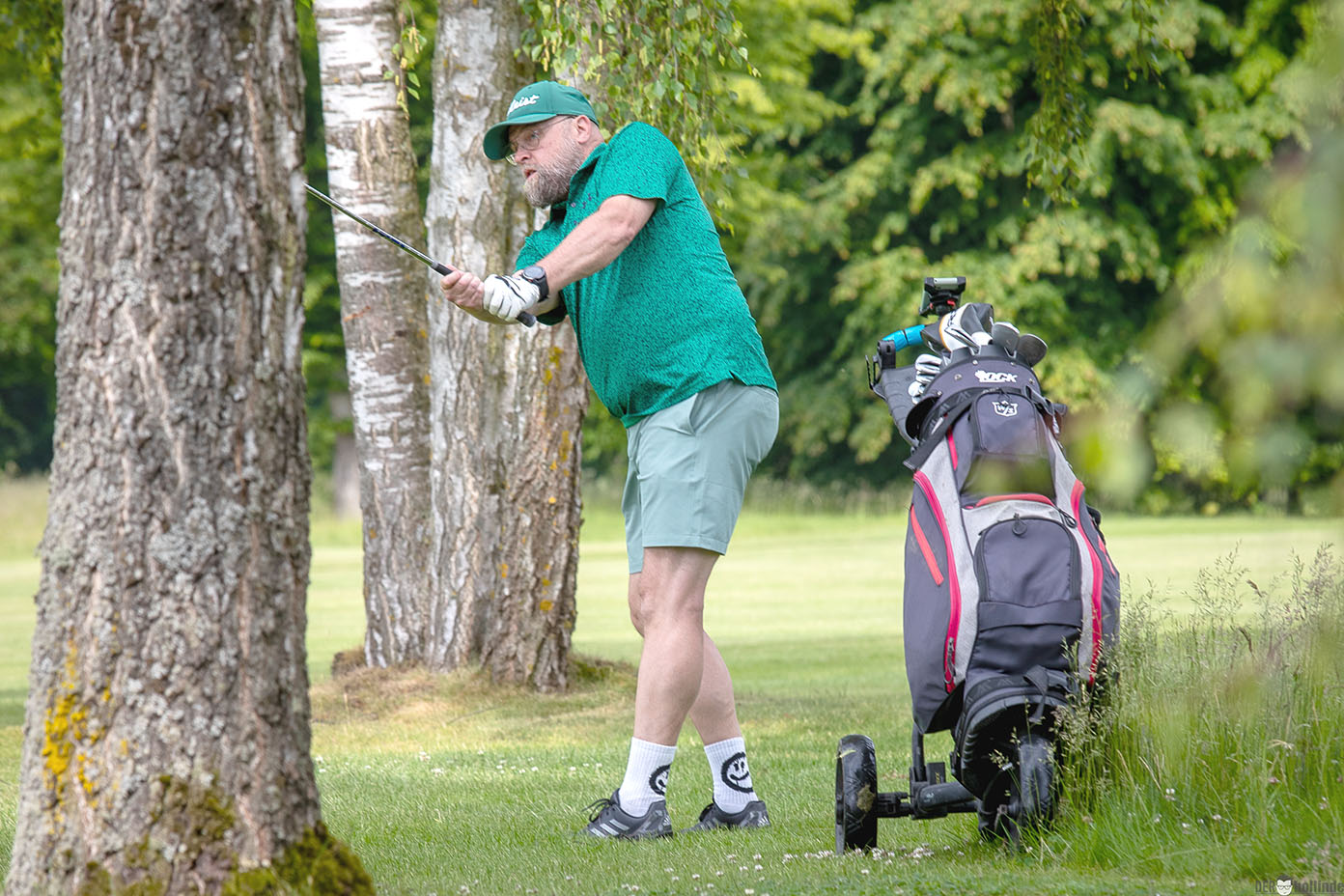 Eine Person in sportlicher Kleidung steht auf einem Golfplatz und bereitet sich darauf vor, einen Golfschlag auszuführen, während ein Golfwagen mit Golftasche neben ihr steht.