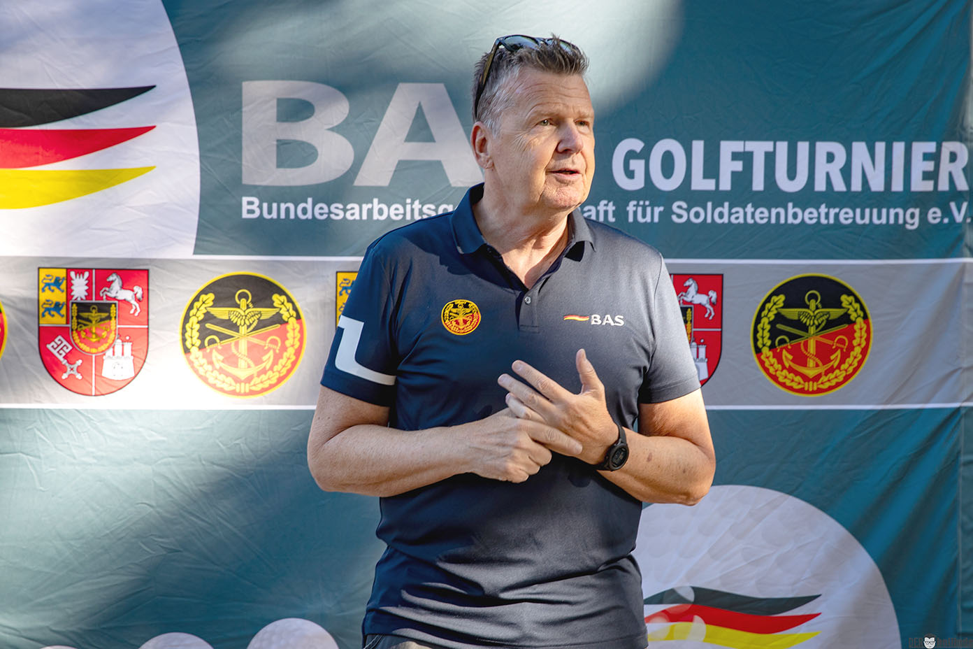 Eine Person mit kurz geschnittenem Haar trägt ein dunkelblaues Polohemd mit Logos und steht vor einem Banner mit deutschen Flaggenfarben und Texten zur Bundesarbeitsgemeinschaft für Soldatenbetreuung bei einem Golfturnier.