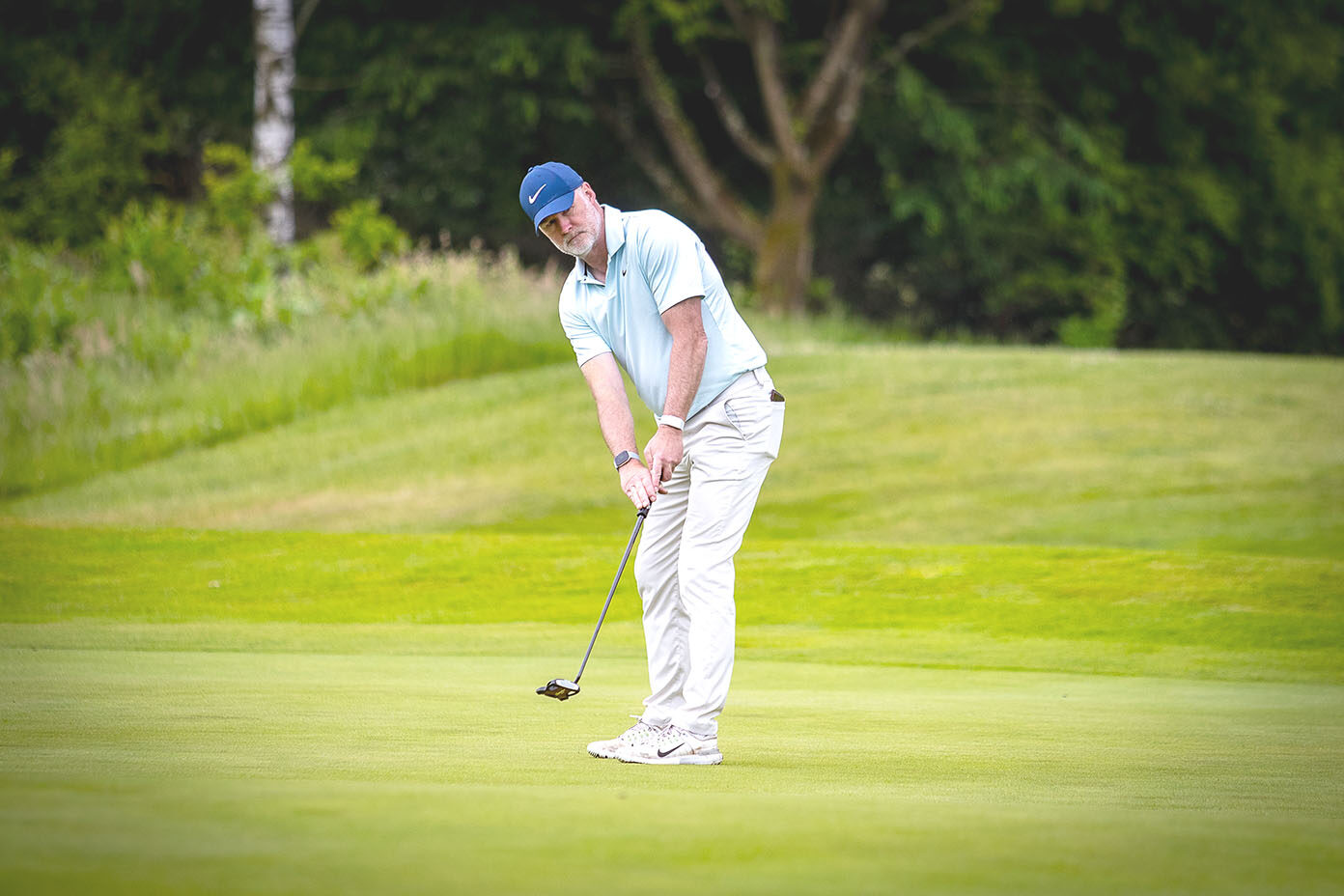 Eine Person trägt ein hellblaues Poloshirt, eine blaue Kappe und helle Hosen und spielt Golf auf einem grünen Golfplatz mit Bäumen im Hintergrund.