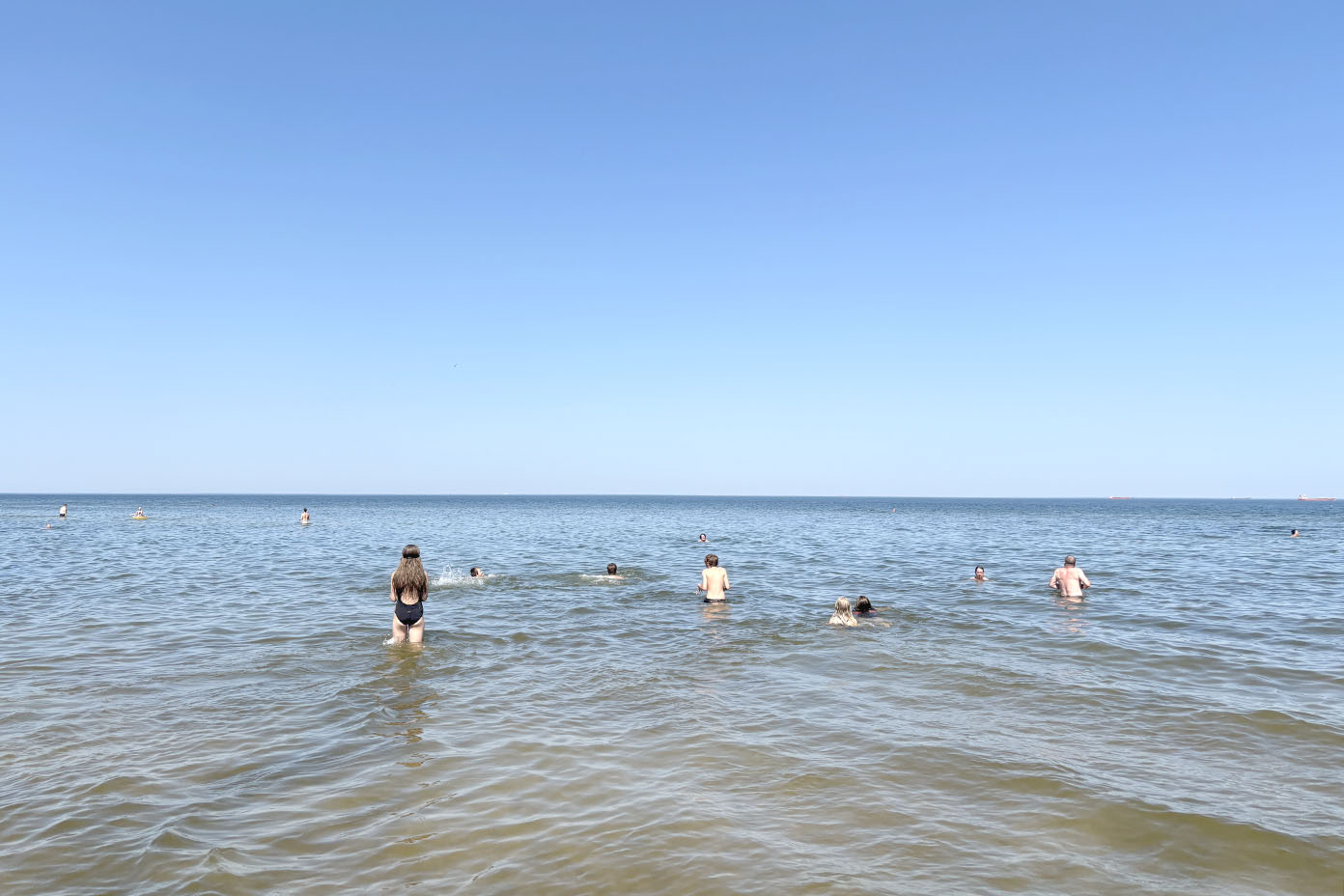 Mehrere Personen schwimmen und stehen im flachen Wasser eines ruhigen Meeres unter klarem blauem Himmel.