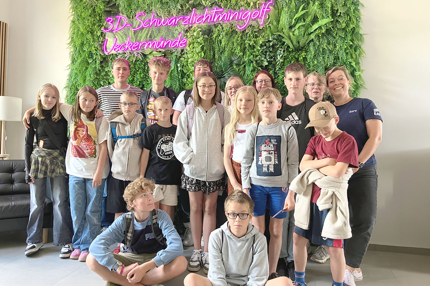 Eine Gruppe von Kindern und Erwachsenen steht vor einer grünen Wand mit Pflanzen und einem leuchtenden Schriftzug in Pink.