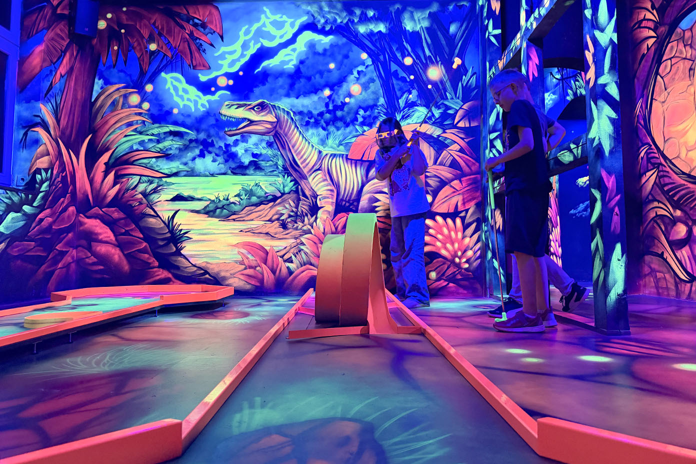 Mehrere Personen spielen Minigolf in einer farbenfroh beleuchteten Halle mit Dinosaurier- und Dschungelmotiven an den Wänden.