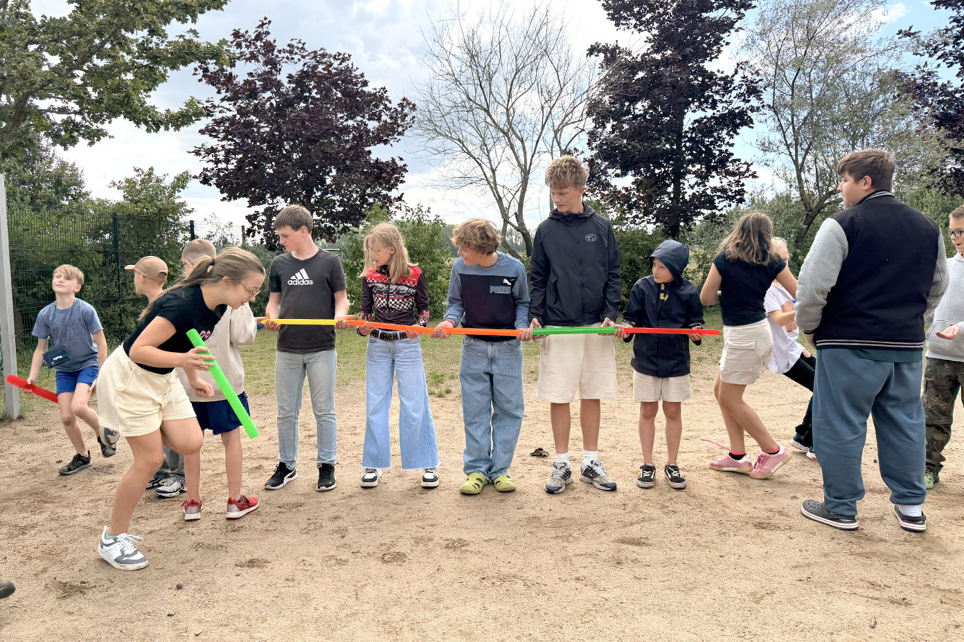 Eine Gruppe von Kindern steht im Freien auf sandigem Boden und hält bunte Stäbe in den Händen, während sie sich auf ein gemeinsames Spiel oder eine Aktivität konzentrieren.