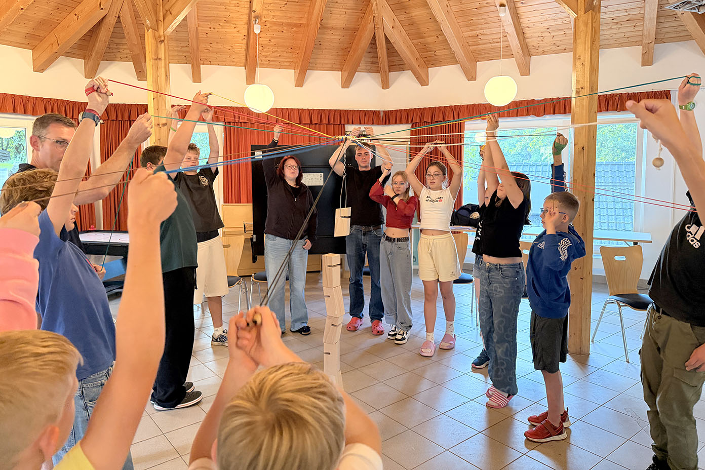 Eine Gruppe von Menschen steht in einem Raum mit Holzdach im Kreis und hält mit ausgestreckten Armen Schnüre, an denen ein Turm aus Holzklötzen hängt.