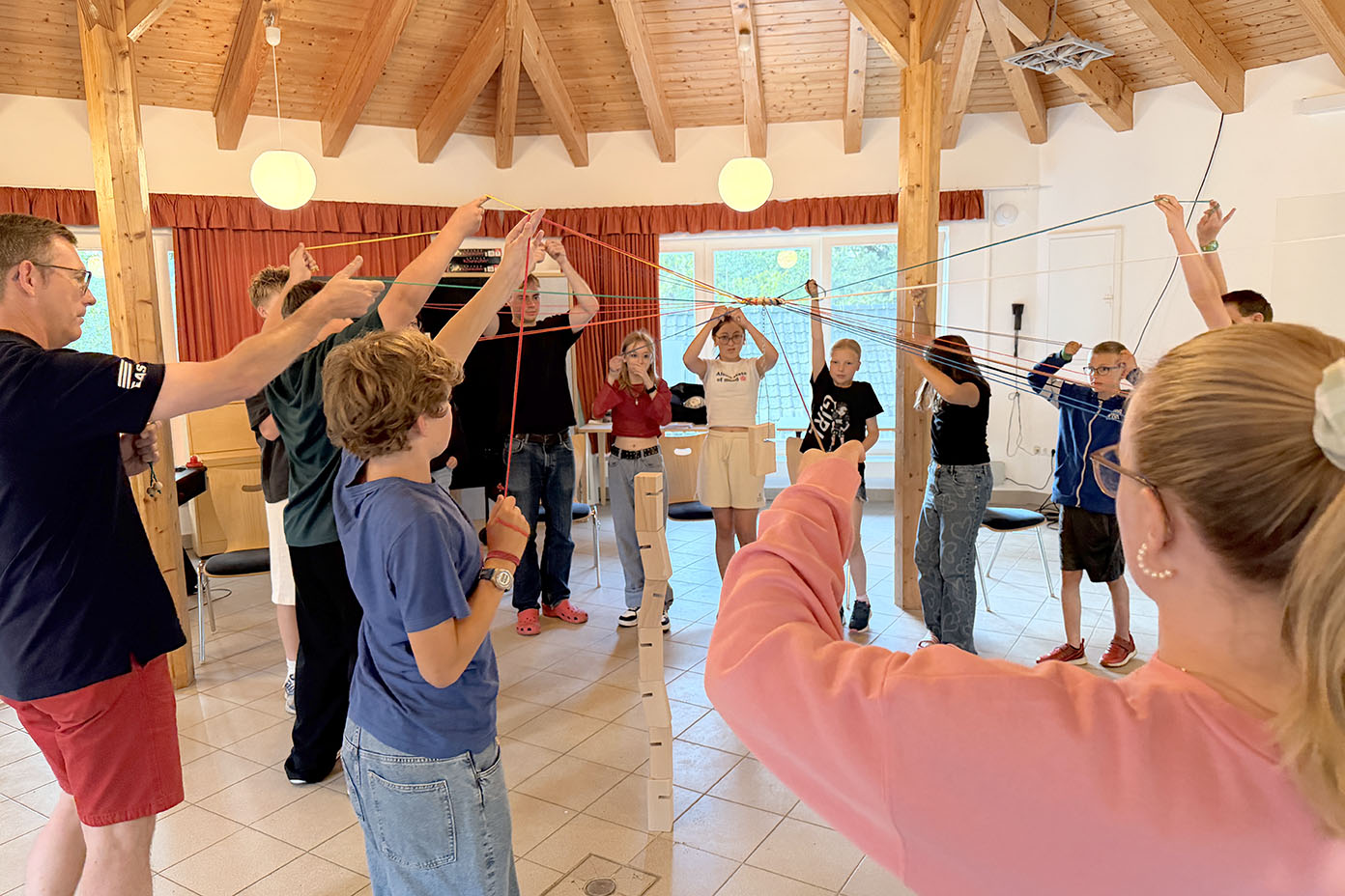 Eine Gruppe von Menschen steht in einem Raum mit Holzbalkendecke im Kreis und hält bunte Fäden, die miteinander verbunden sind, als Teil einer Team- oder Kommunikationsübung.