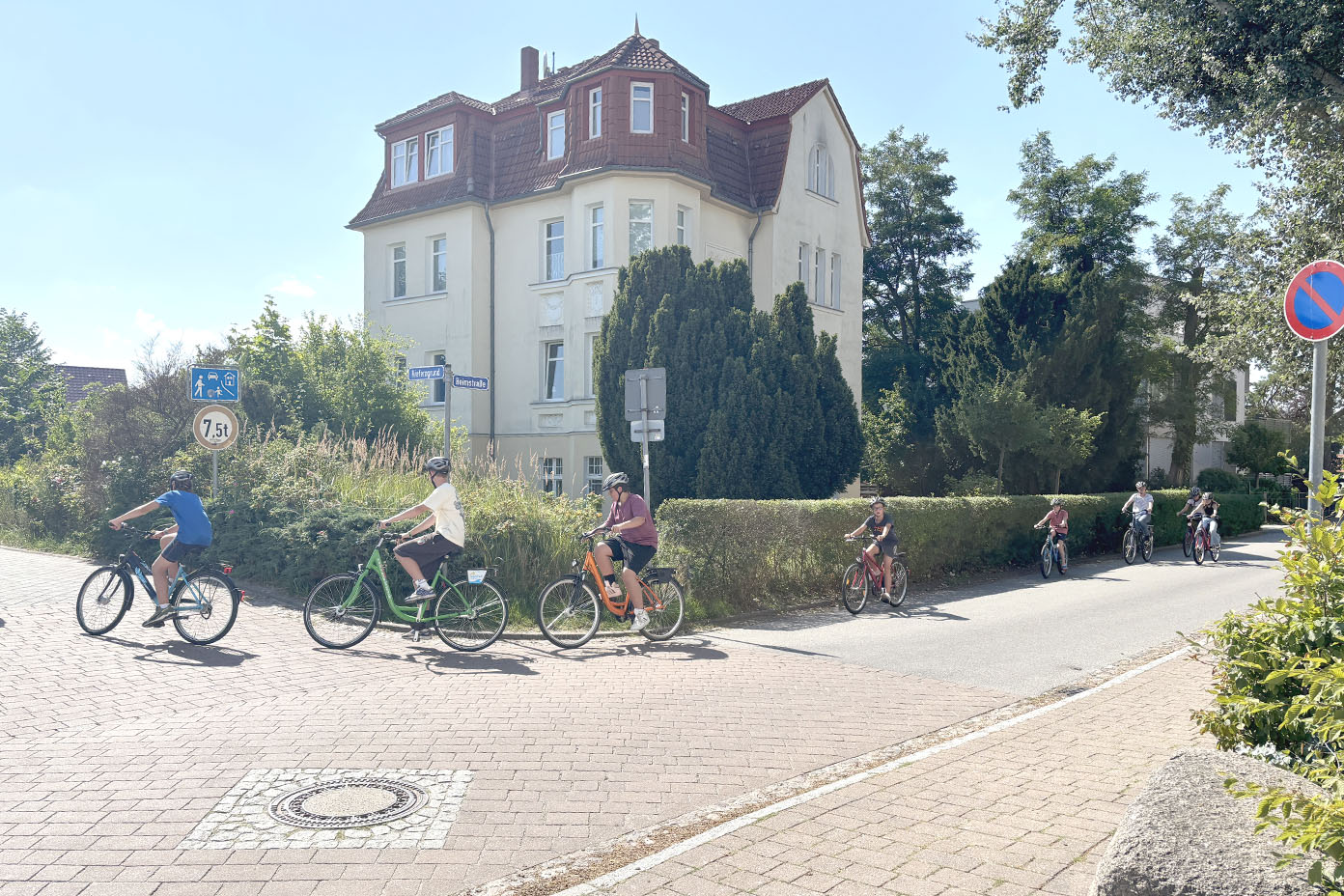 Mehrere Personen fahren bei sonnigem Wetter mit Fahrrädern auf einem gepflasterten Weg an einem großen weißen Haus mit rotem Dach und viel Grün entlang.