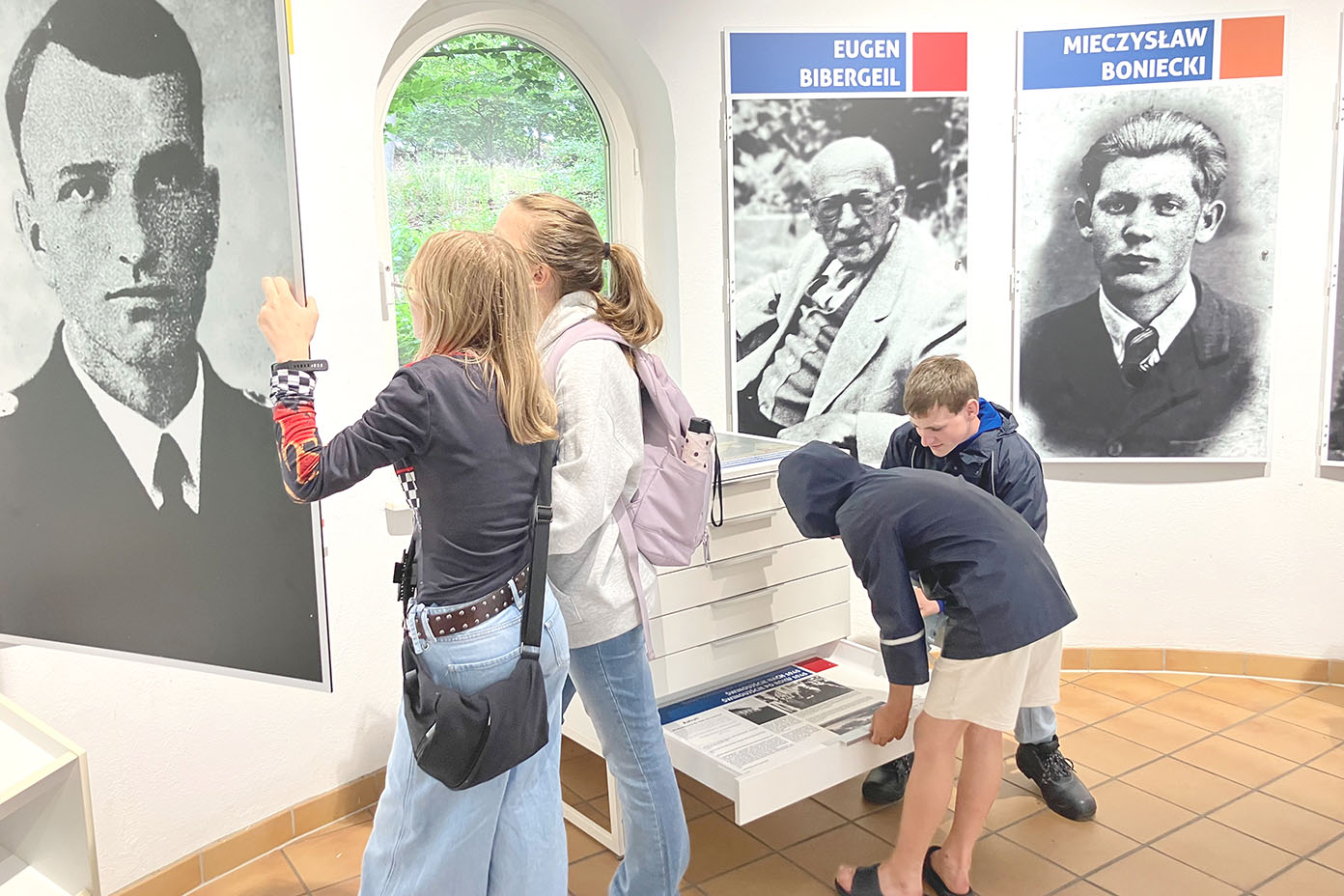 Mehrere Personen betrachten in einem Ausstellungsraum große Schwarz-Weiß-Porträtfotos und lesen Informationen von einer Ausstellungstafel.