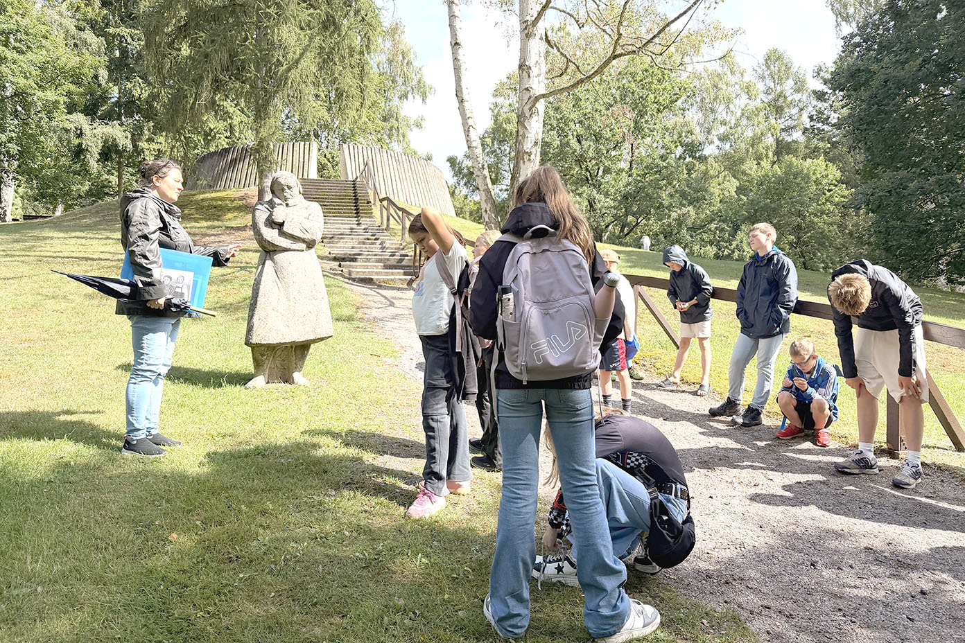 Eine Gruppe von Menschen steht und kniet auf einem Weg in einem Park neben einer Statue einer stehenden Person, umgeben von Bäumen und Gras.