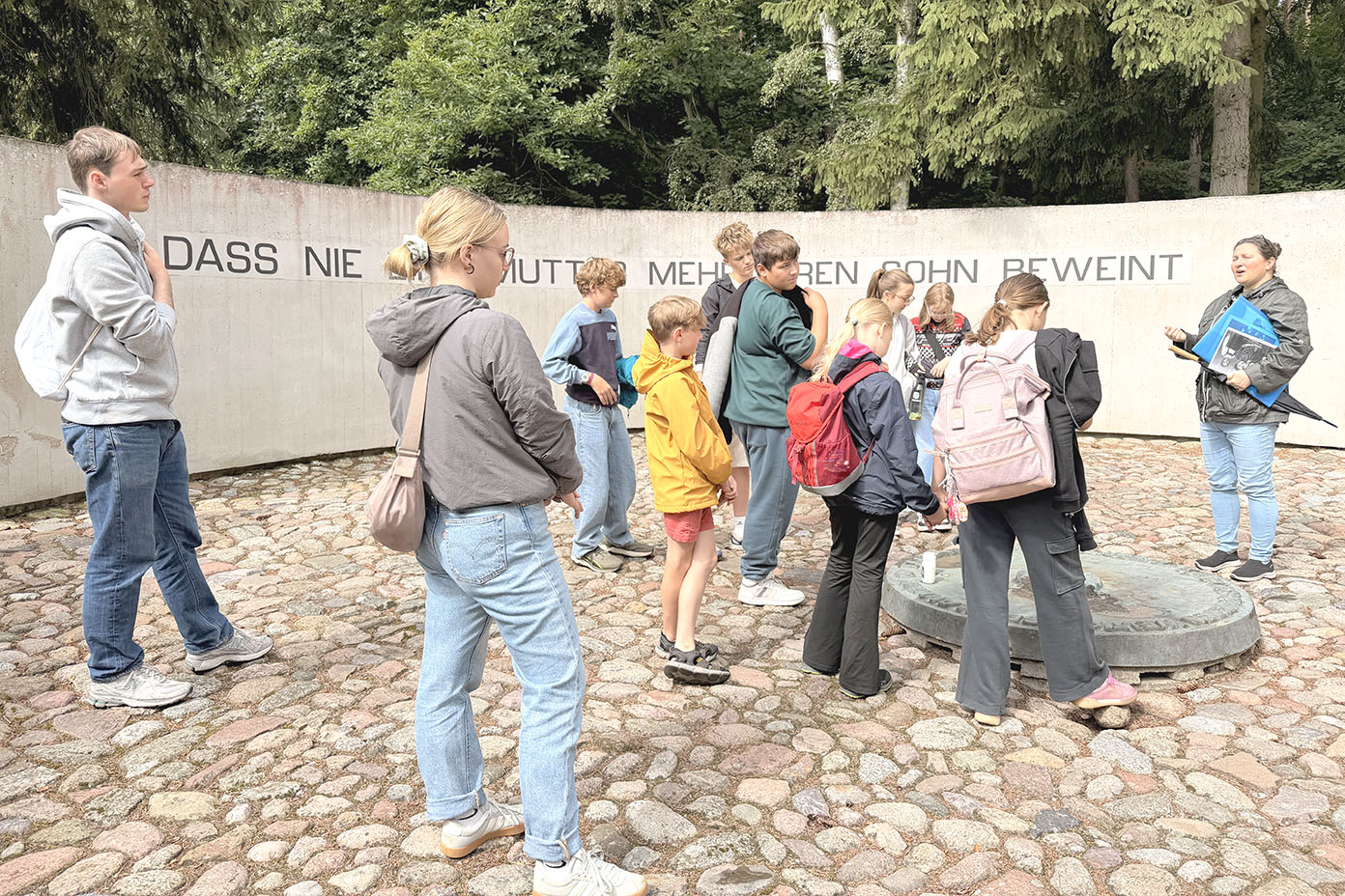 Eine Gruppe von Menschen steht auf einem gepflasterten Platz vor einer halbkreisförmigen Wand mit deutscher Schrift und hört einer Person zu, die etwas erklärt.