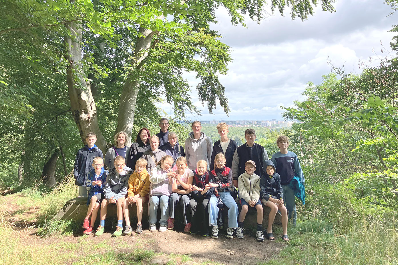 Eine Gruppe von Menschen sitzt und steht auf einem Waldweg unter Bäumen bei sonnigem Wetter mit Blick auf eine Stadt im Hintergrund.