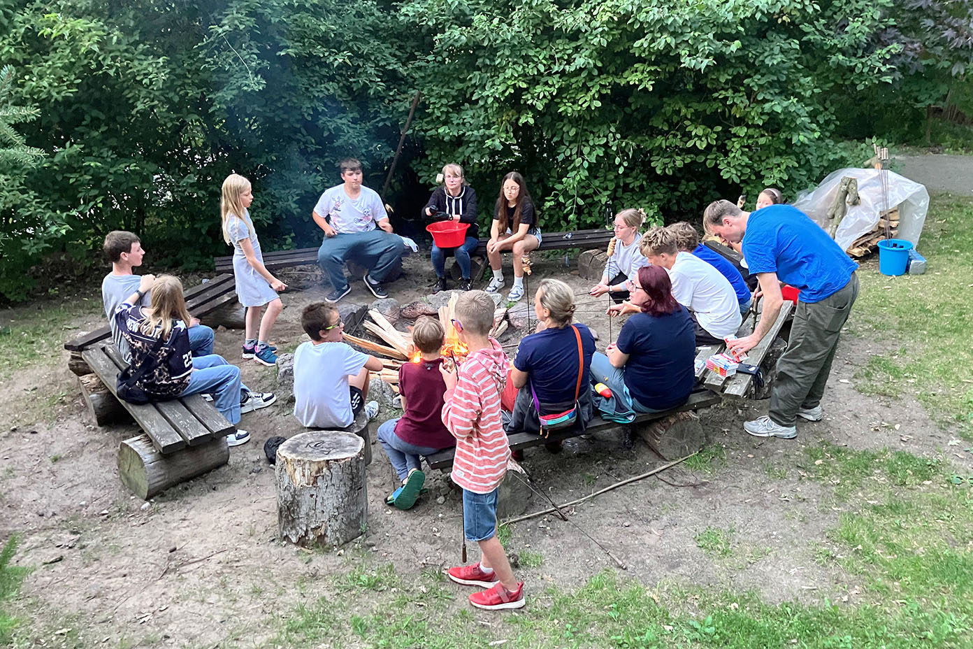 Eine Gruppe von Kindern und Erwachsenen sitzt und steht um ein Lagerfeuer im Freien, umgeben von Bäumen, während sie sich unterhalten und Stöcke ins Feuer halten.