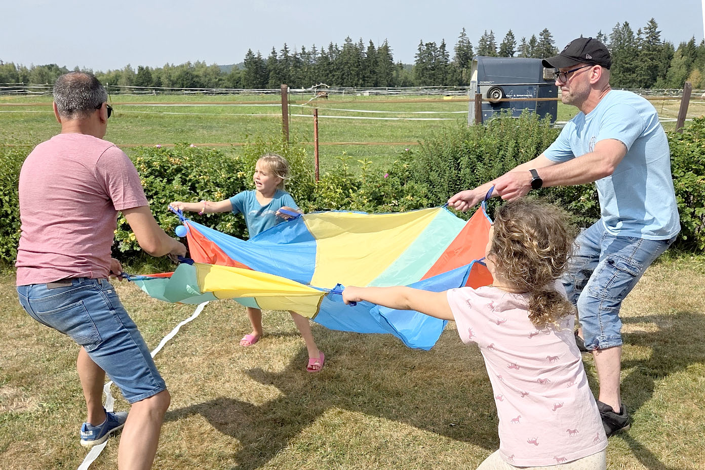 Vier Personen, darunter zwei Kinder, spielen gemeinsam mit einem bunten Tuch auf einer Wiese an einem sonnigen Tag.