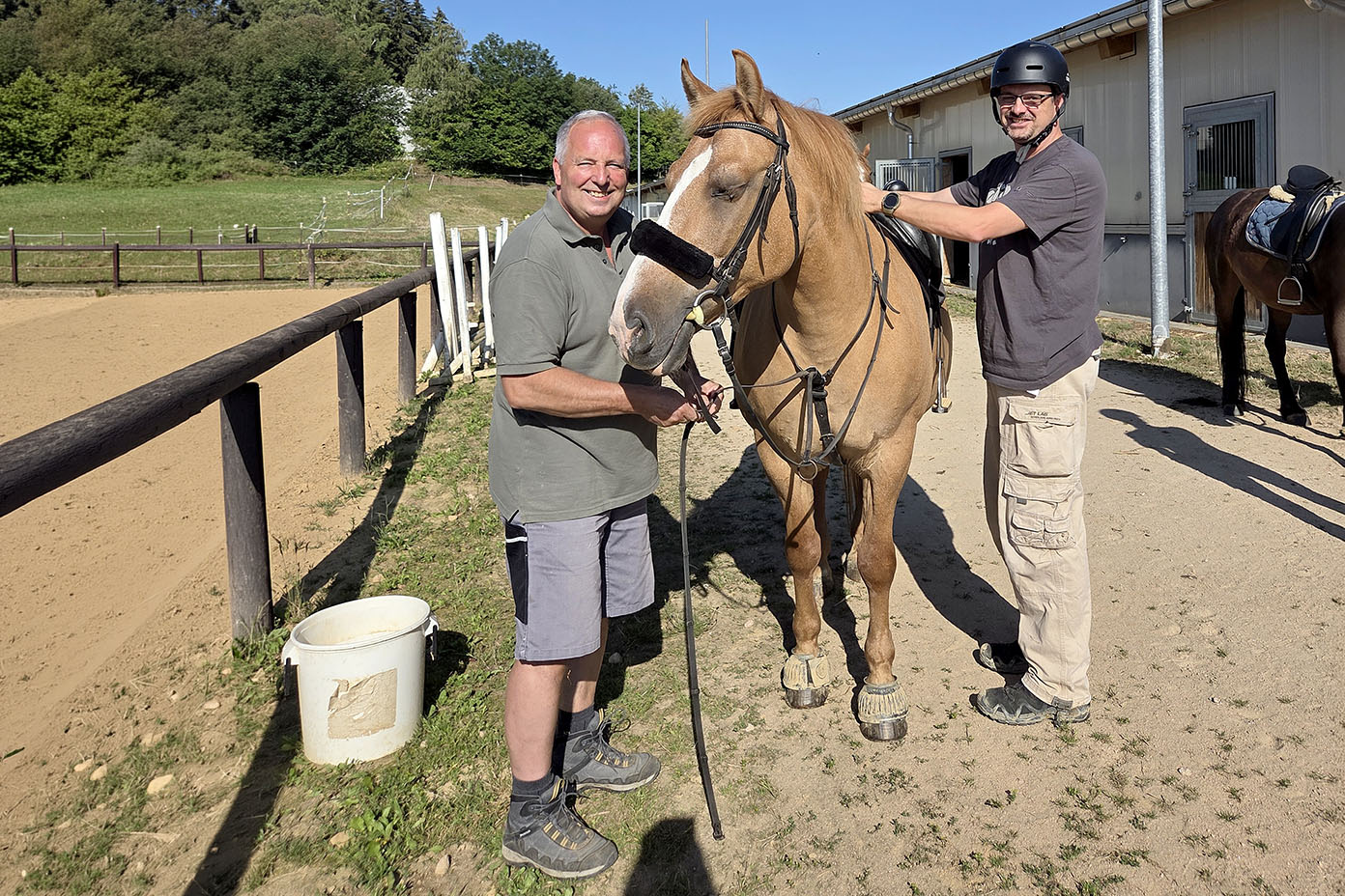 Zwei Personen stehen neben einem braunen Pferd auf einem Reitplatz, eine trägt einen Helm und hält die Zügel, die andere steht vorne am Pferd und hält es am Halfter.