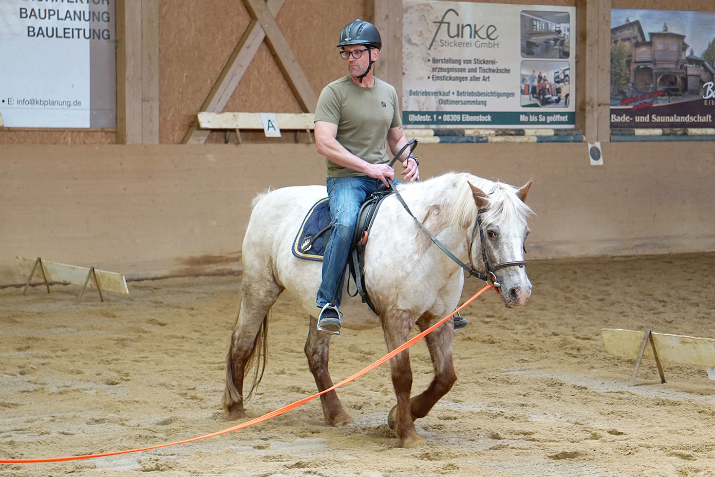 Eine Person mit Helm reitet auf einem weißen Pferd in einer Reithalle mit Sandboden und Werbetafeln an der Wand.
