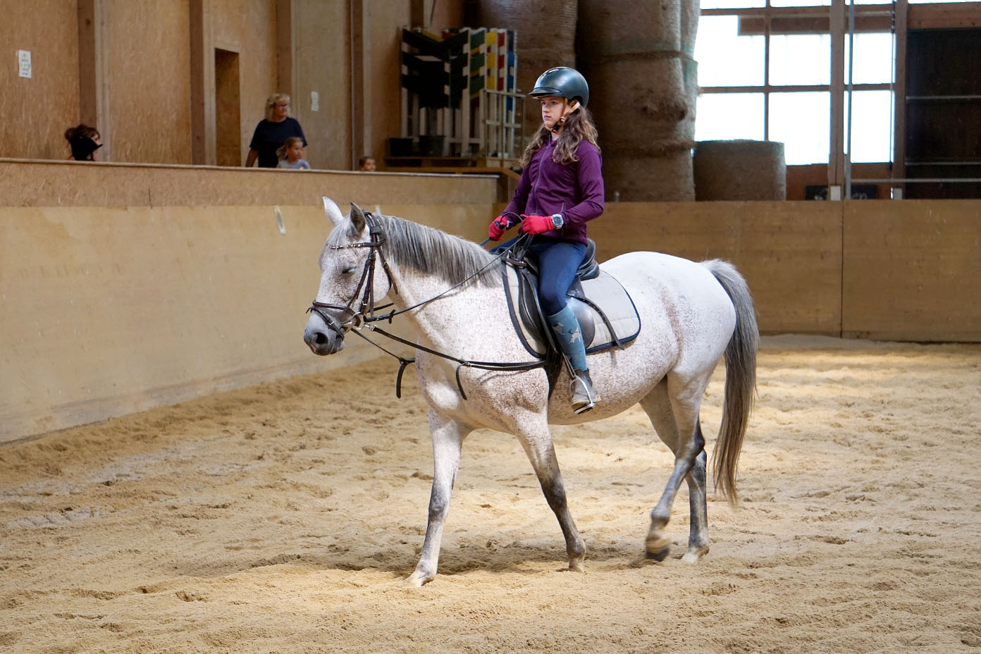 Eine Person mit Helm und Reitkleidung sitzt auf einem weißen Pferd in einer Reithalle mit Sandboden.
