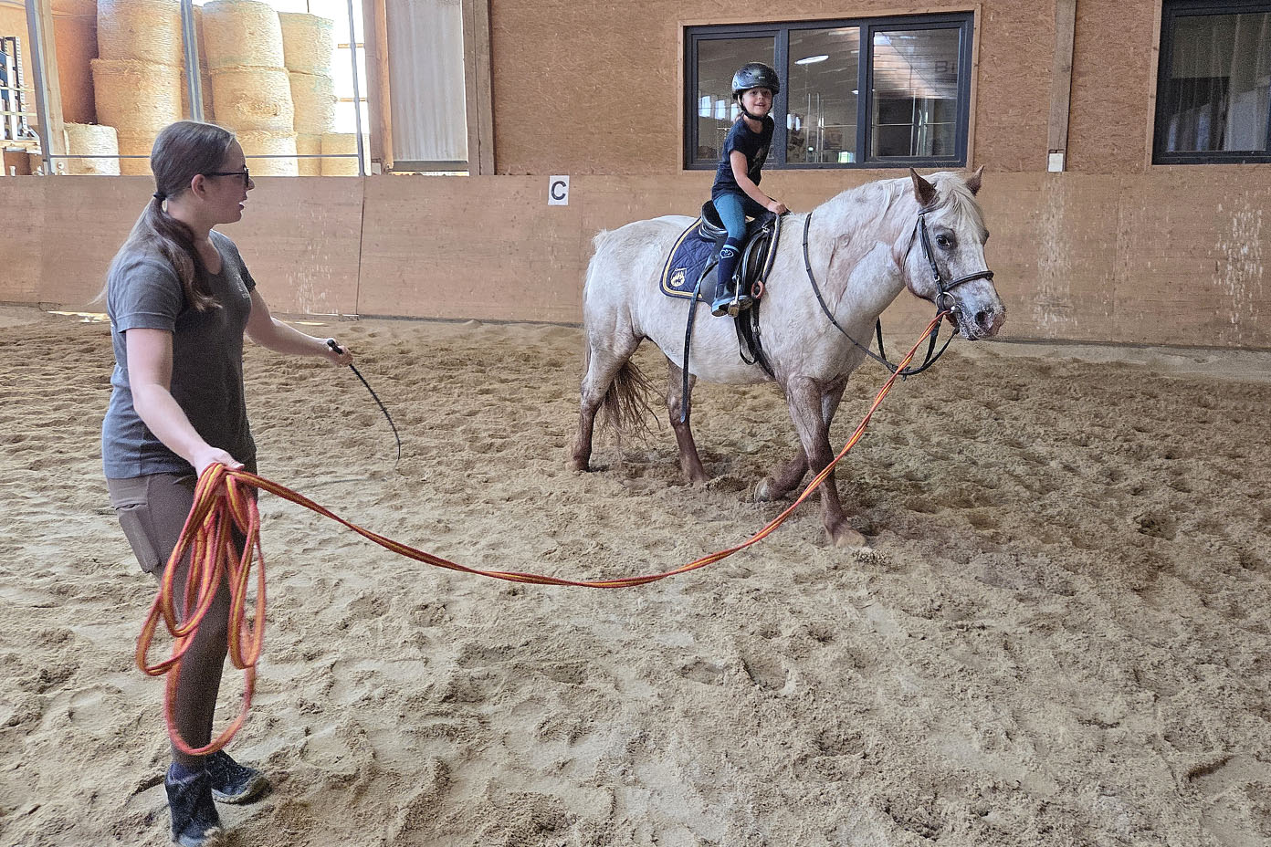 Eine Person hält ein langes Seil und führt ein Pferd, auf dem ein Kind mit Helm sitzt, in einer Reithalle.