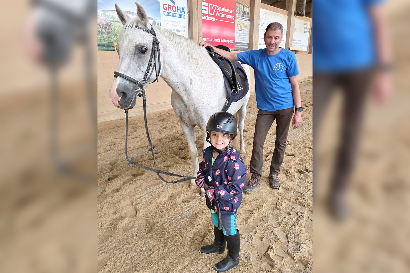 Ein Kind mit Helm und Gummistiefeln hält die Zügel eines weißen Pferdes, während eine Person in blauer Kleidung das Pferd im Innenbereich eines Reitstalls hält.