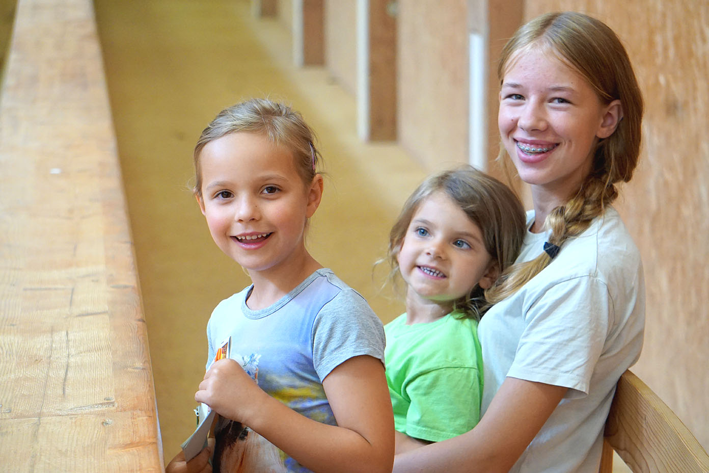Drei Kinder sitzen lächelnd nebeneinander auf einer Bank in einem hellen Flur mit Holzwänden.