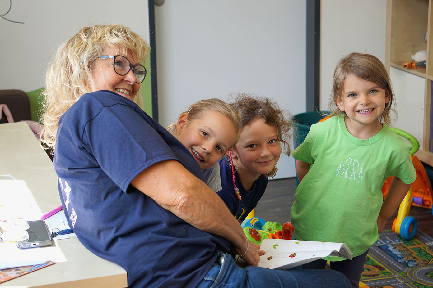 Eine Person mit Brille und blauem T-Shirt sitzt an einem Tisch und umarmt drei lächelnde Kinder, die in einem hellen Raum mit Spielzeug stehen.
