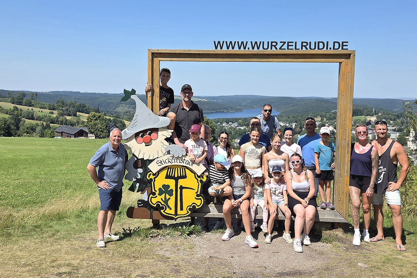Eine Gruppe von Menschen posiert bei sonnigem Wetter vor einer großen Holzrahmenstruktur mit der Aufschrift www.wurzelrudi.de und einer bunten Cartoonfigur in einer grünen Landschaft mit Hügeln und einem See im Hintergrund.