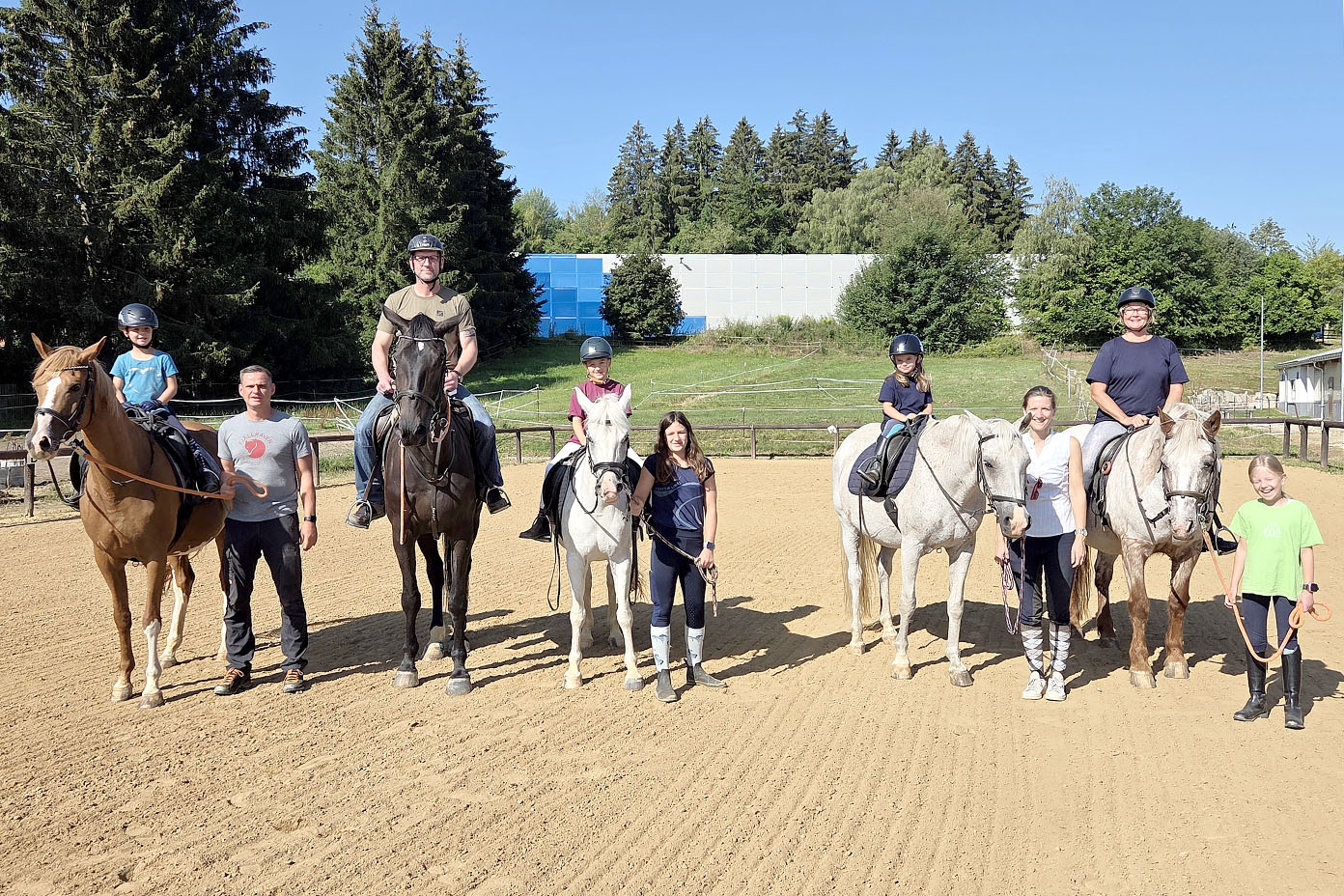 Mehrere Personen stehen und sitzen auf Pferden auf einem Reitplatz bei sonnigem Wetter, umgeben von Bäumen und einer Wiese.