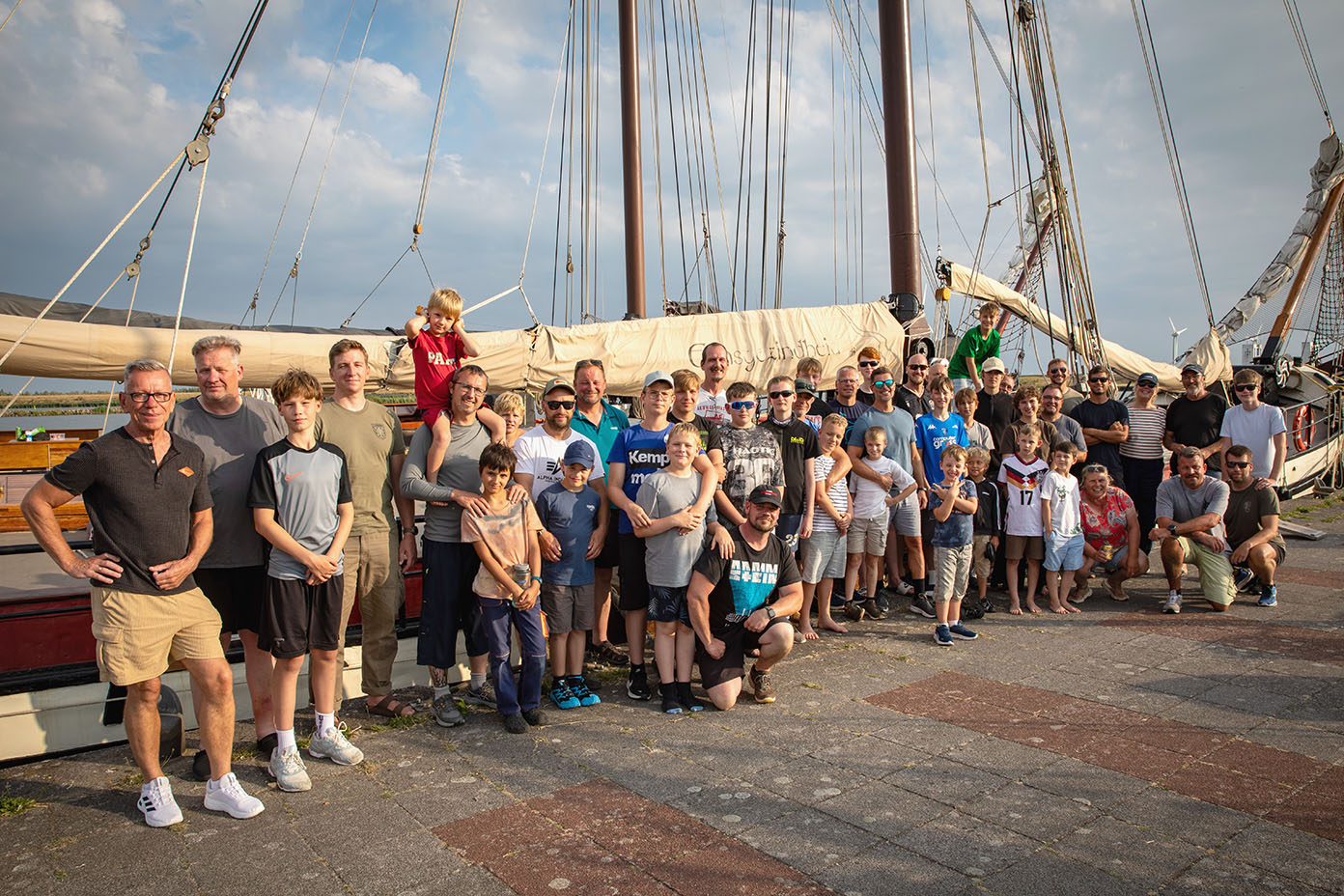 Eine große Gruppe von Menschen unterschiedlichen Alters steht bei sonnigem Wetter vor einem Segelschiff mit hohen Masten und Segeln.