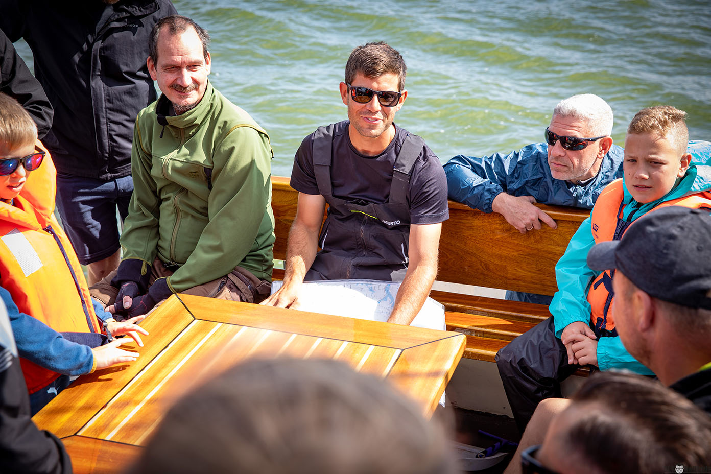 Mehrere Personen sitzen auf einem Boot bei sonnigem Wetter, einige tragen Sonnenbrillen und Rettungswesten, während sie sich um einen Holztisch versammeln.