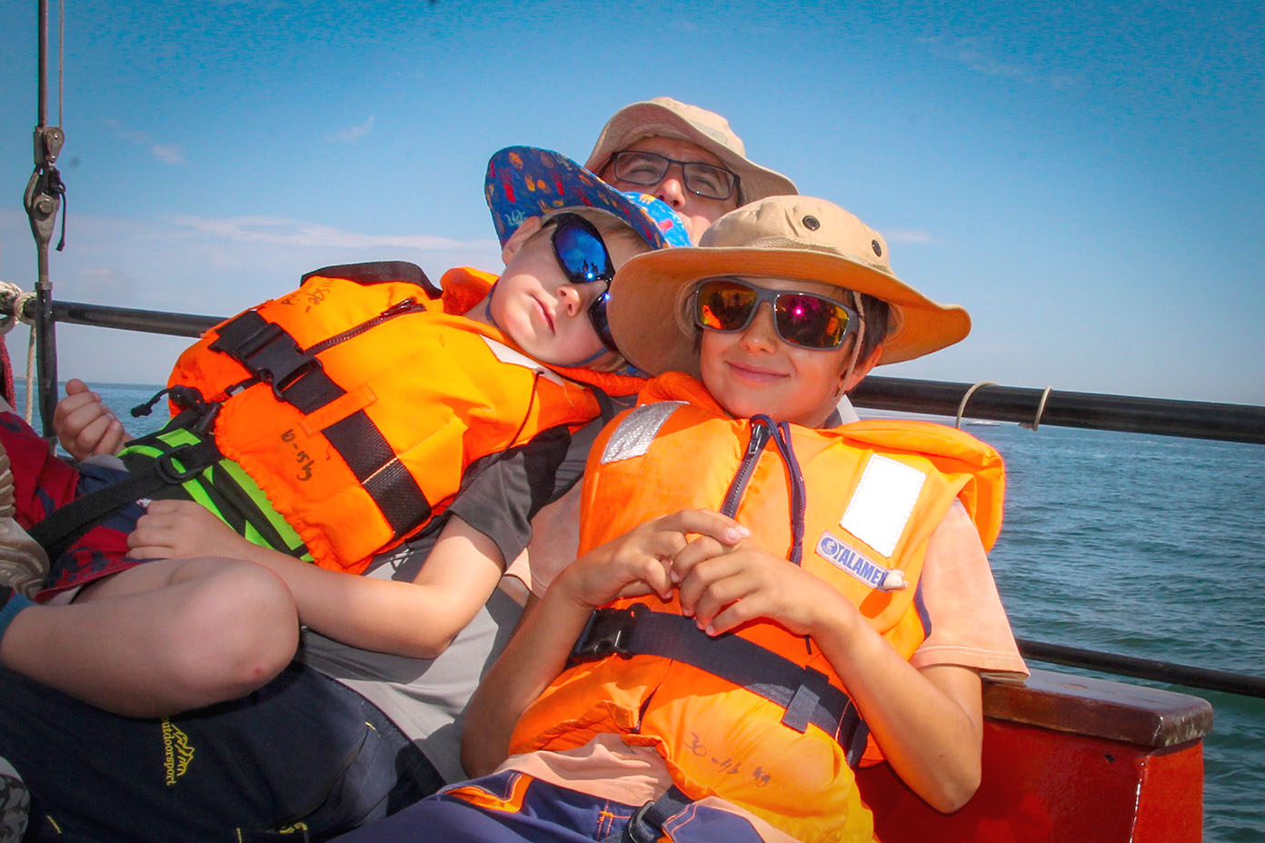 Drei Personen tragen bunte Hüte und orangefarbene Schwimmwesten und sitzen entspannt auf einem Boot bei sonnigem Wetter auf dem Wasser.