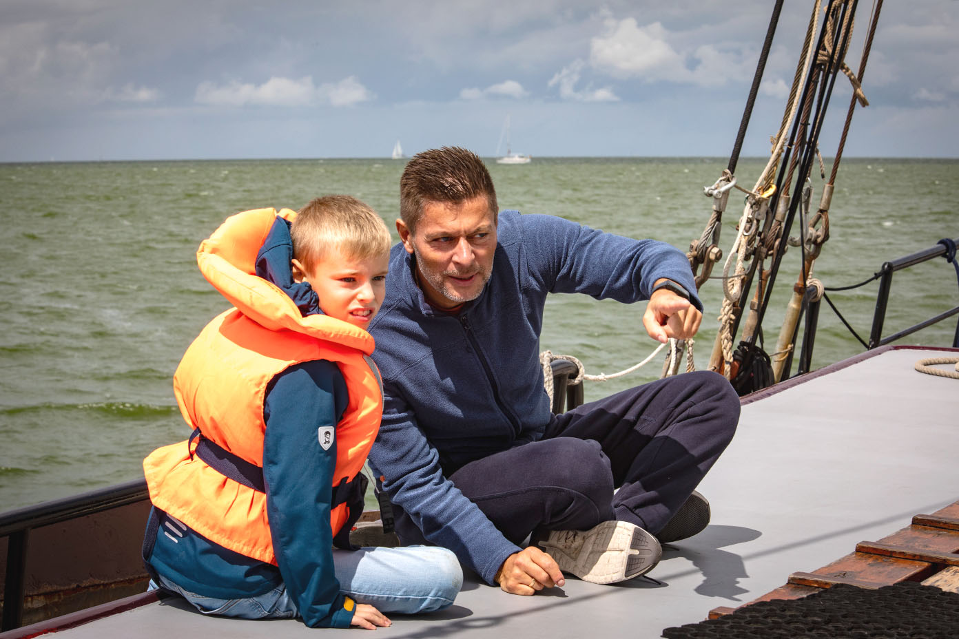 Ein Kind mit einer orangen Rettungsweste sitzt neben einer anderen Person in Freizeitkleidung auf dem Deck eines Segelbootes auf offenem Wasser.