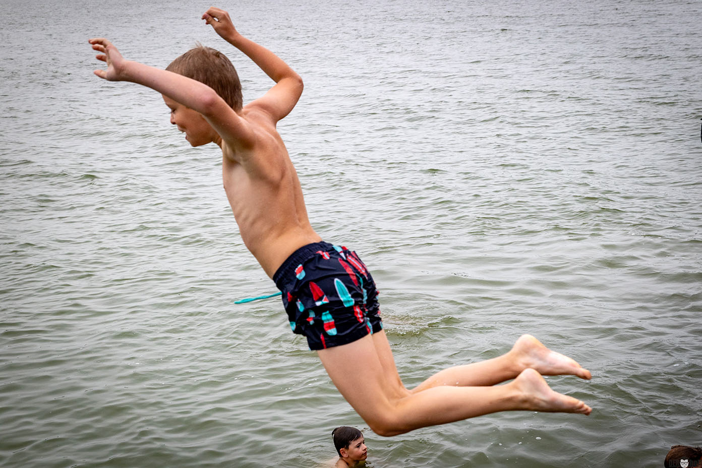 Ein Kind mit kurzen Haaren und bunten Badehosen springt mit ausgestreckten Armen in ein Gewässer, während eine weitere Person im Wasser zu sehen ist.