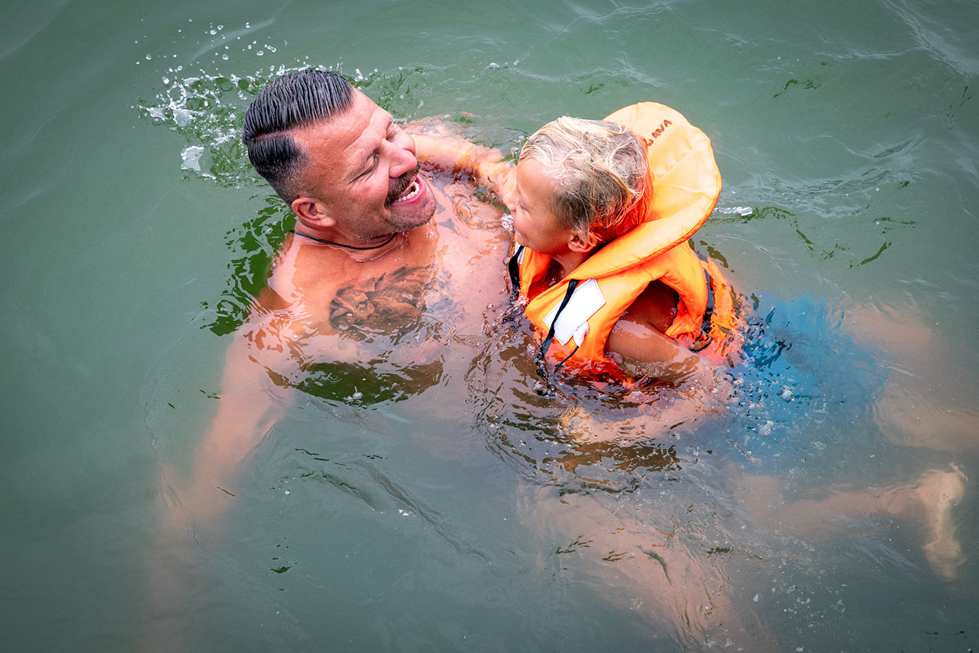 Eine Person mit kurzen Haaren und ein Kind mit Schwimmweste lachen und spielen gemeinsam im Wasser.