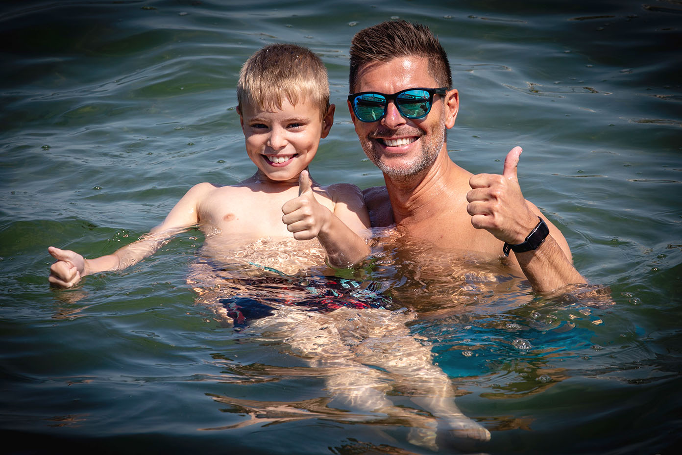 Ein Kind und ein Erwachsener mit Sonnenbrille schwimmen im Wasser und zeigen beide einen Daumen nach oben, während sie lächeln.