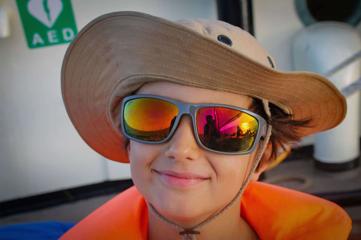 Eine Kind trägt einen breitkrempigen Hut, eine orangefarbene Schwimmweste und reflektierende Sonnenbrillen, in denen eine weitere Person und eine bunte Umgebung zu sehen sind.