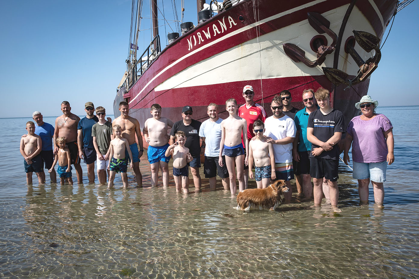 Eine Gruppe von Menschen unterschiedlichen Alters steht im flachen Wasser vor einem großen Segelschiff mit dem Namen Nirvana an einem sonnigen Tag.