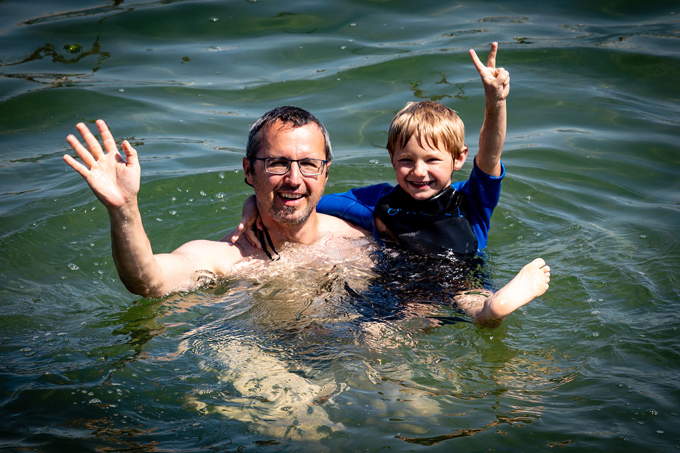 Eine Person hält ein lachendes Kind im Wasser, beide winken mit den Händen und wirken fröhlich.