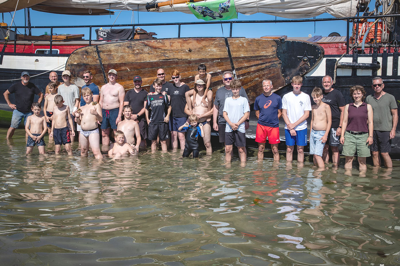 Eine Gruppe von Menschen steht knietief im Wasser vor einem großen Segelschiff bei sonnigem Wetter.