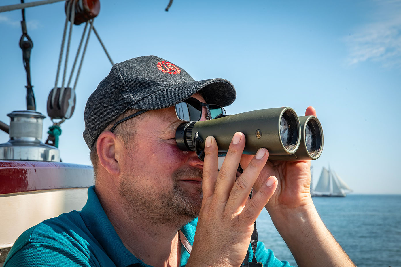 Eine Person mit Kappe schaut durch ein Fernglas auf einem Boot bei klarem Himmel und ruhigem Meer, im Hintergrund ist ein Segelboot zu sehen.