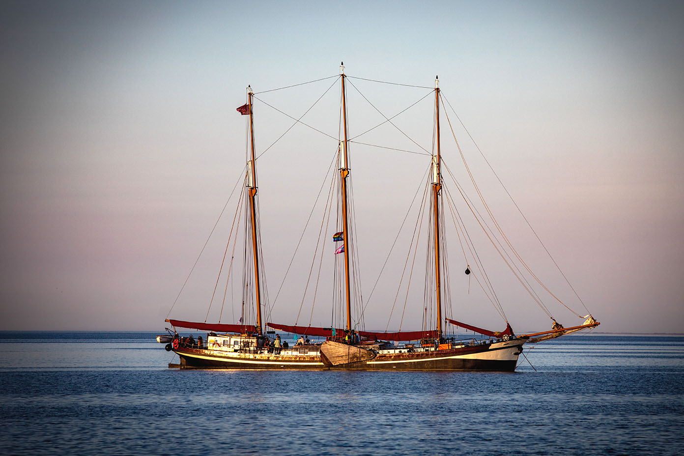 Ein Segelschiff mit drei Masten liegt ruhig auf dem Wasser unter einem klaren Himmel.