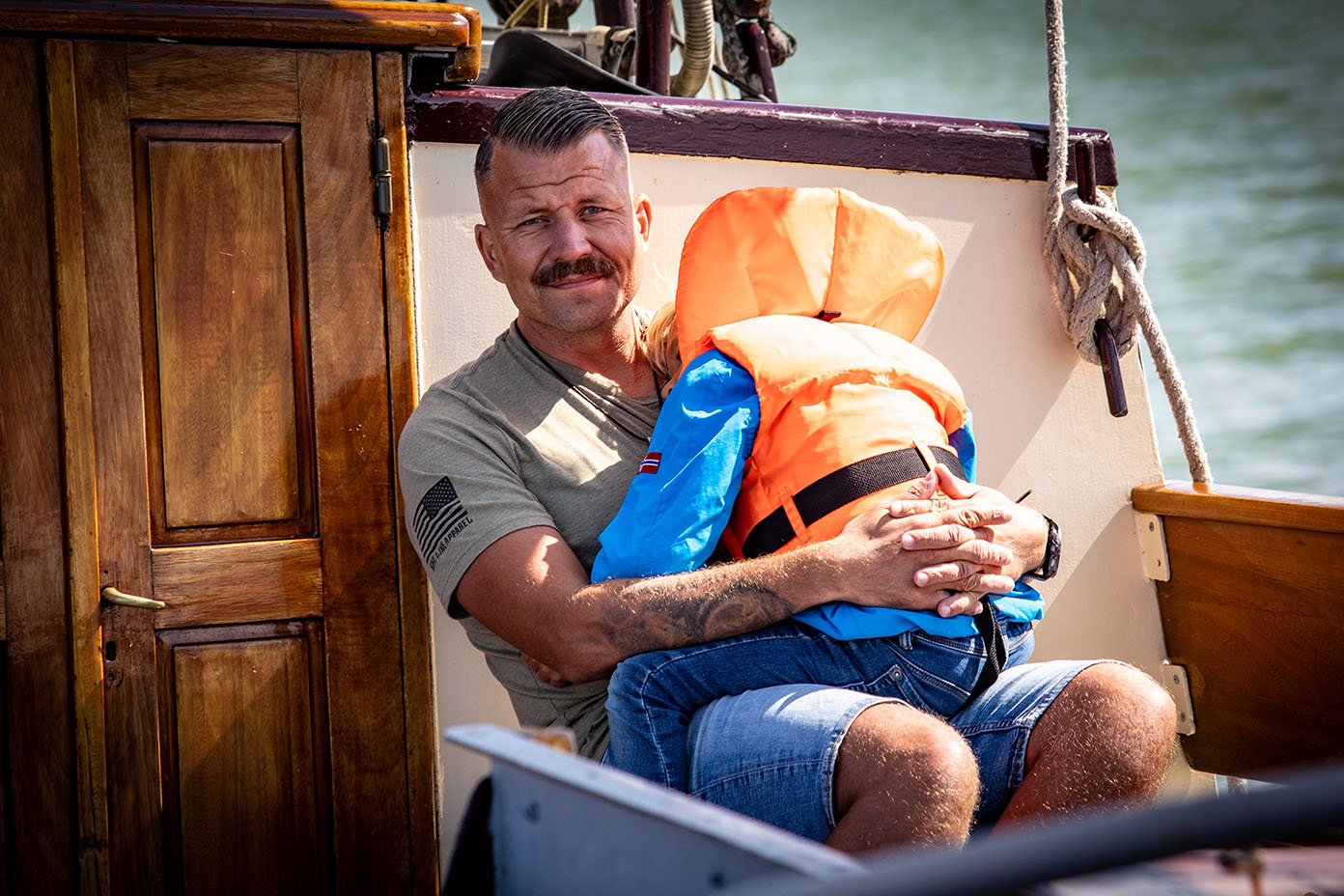 Eine Person mit kurzem Haar und Schnurrbart sitzt auf einem Boot und hält ein Kind mit orangefarbener Schwimmweste und blauer Kleidung im Arm.