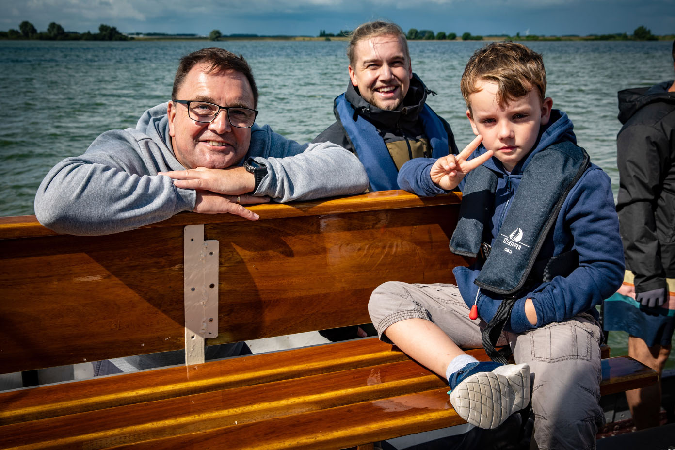 Drei Personen mit Rettungswesten sitzen auf einer Bank an einem Gewässer, wobei eine Person ein Peace-Zeichen mit den Fingern zeigt.