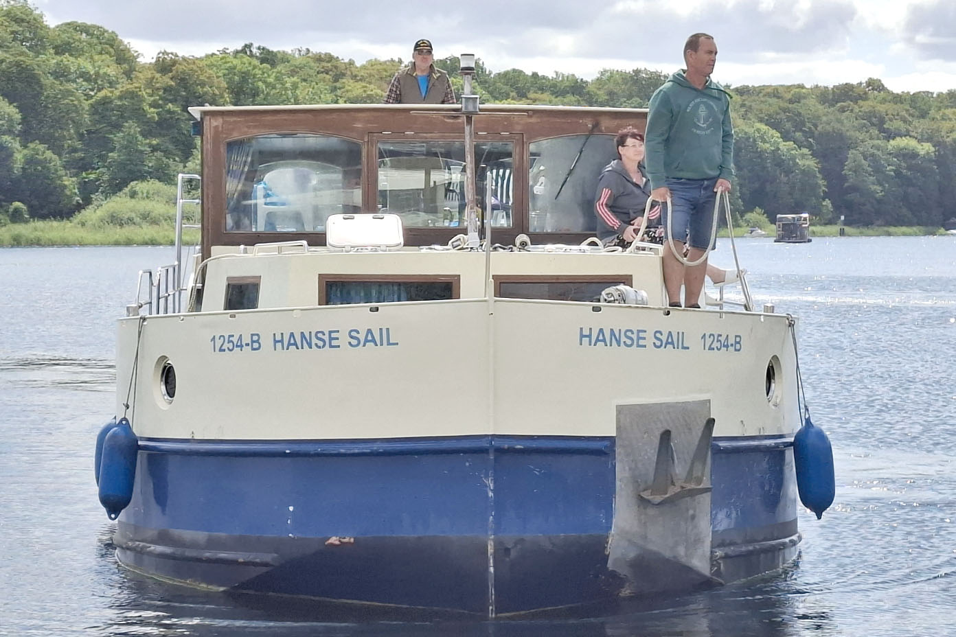 Ein Boot mit der Aufschrift Hanse Sail fährt auf einem See, auf dem Deck stehen drei Personen, umgeben von grünen Bäumen am Ufer.