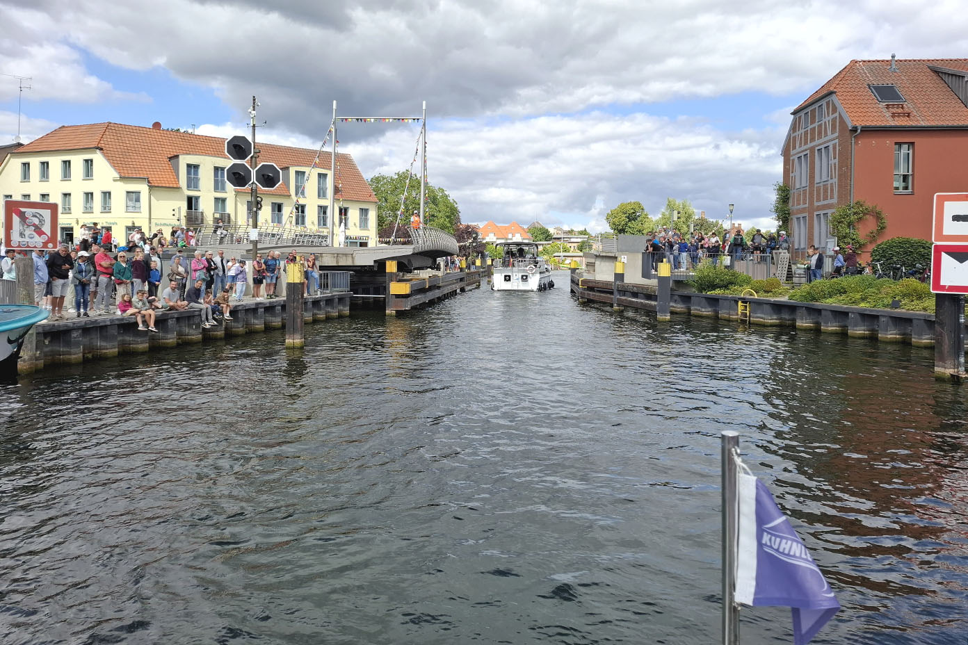 Ein Kanal mit einem Boot in der Mitte und vielen Menschen, die an beiden Uferseiten auf Stegen und vor Gebäuden stehen, unter einem bewölkten Himmel.