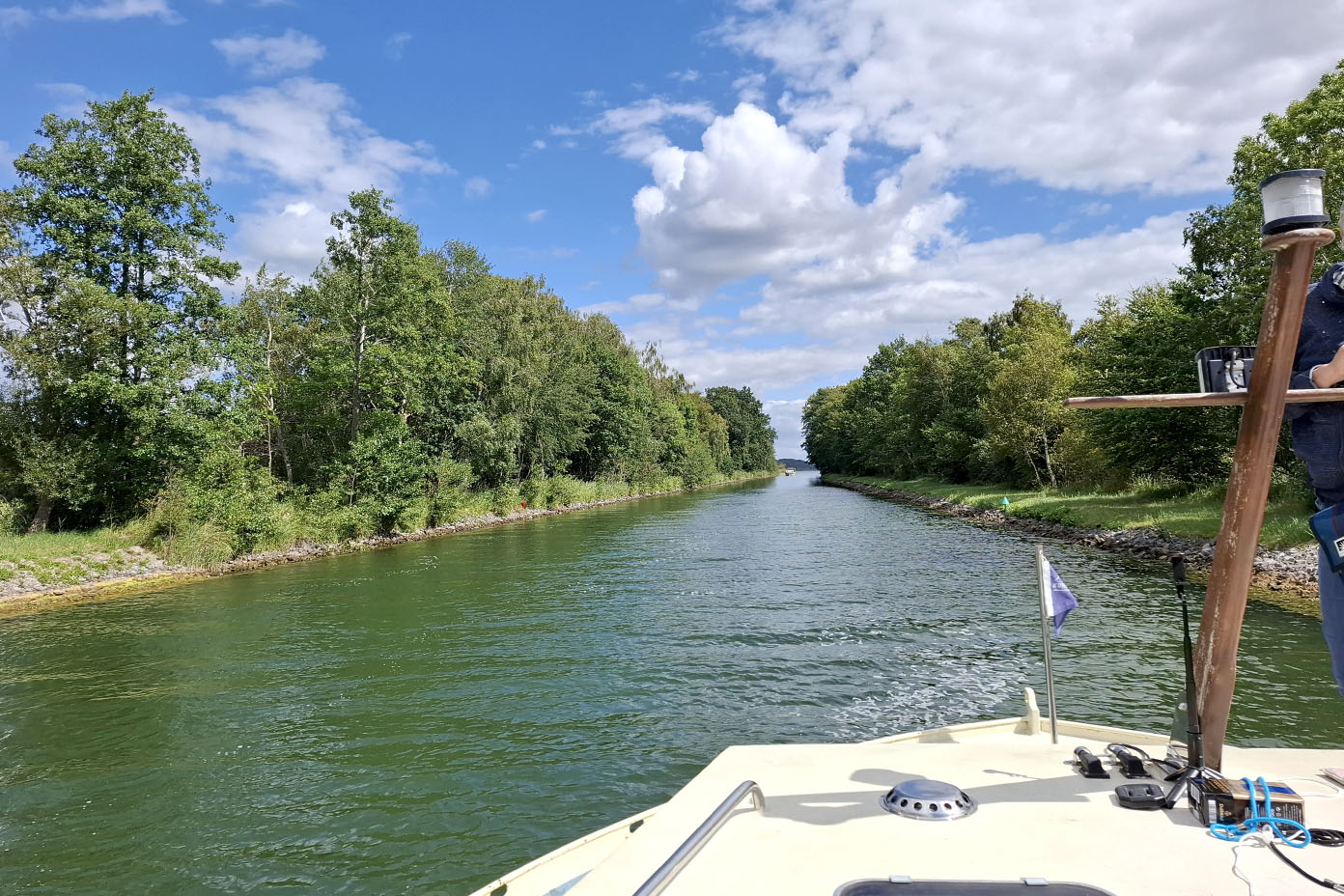Ein Boot fährt auf einem ruhigen Fluss oder Kanal, umgeben von grünen Bäumen unter einem teilweise bewölkten Himmel.